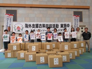 写真：児童とボランティアの人が集まった衣料の前で「世界の友達に届けてね」書かれたカードを持って記念写真を撮る様子