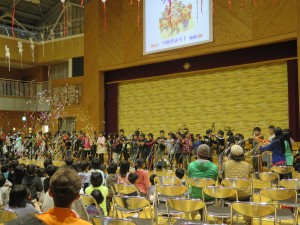 写真：大安中学校の体育館で」ブラスバンドの演奏する児童と演奏を聴く参加者