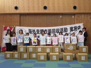 写真:世界の子ども届けてねと一字一字書かれた紙を持った子どもたちと手分けして箱に入れた衣料と記念撮影