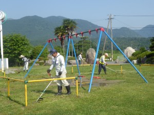 写真：石榑東自治会長OBの方が石榑小学校の校庭にあるブランコの周りの草を草刈り機で刈り取る様子